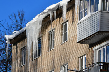 Big beautiful icicles hang from the snow-covered roof in front of the windows of the house. Snow on the roof. Frosty sunny day