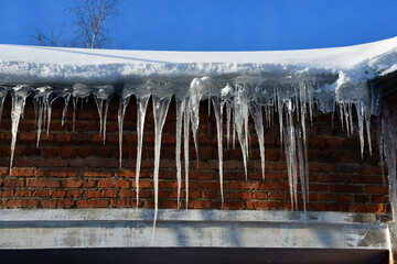 Big beautiful icicles hang from the snow-covered roof of the house. shadow on the wall. Snow on the roof. Frosty sunny day