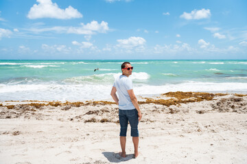 Young happy man smiling standing on sand sandy beach in Miami, Florida with turquoise blue green ocean sea waves background of flying pelican bird