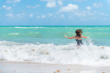 Back of young woman in swimsuit with waves splashing crashing in Sunny Isles, Miami Beach, Florida with Atlantic ocean sea green turquoise water and sargassum seaweed