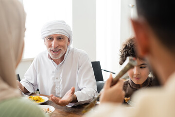smiling muslim man pointing with hand during dinner with arabian grandson and blurred family.