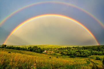 Full arc of double rainbow over summer evening rural landscape - panoramic view of hills with deep forests covered, golden colored meadow with sun beams illuminated, village near forest