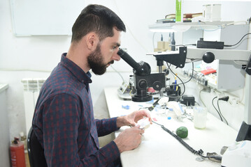 A young man looks through a microscope examining repairs dentures or a jaw in the workshop of a dental technician