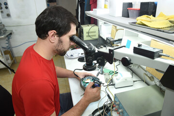 A young man in the workshop examines the microcircuit of an electrical device through a microscope,...