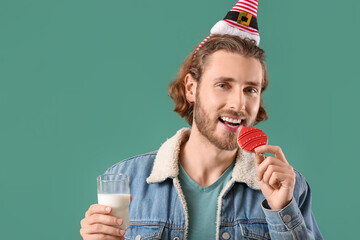 Handsome man with glass of milk eating gingerbread cookie on green background