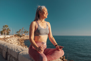 Sporty woman in sports clothes sitting by the seashore, holding buttle of water and mobile phone, looking at the sun