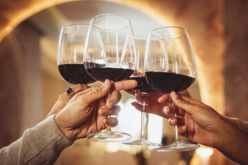 Four friends clinking wineglasses with red wine at restaurant - closeup shot focus on the hands holding glasses and toasting