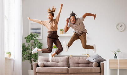 Young energetic active african american family couple jumping on sofa, having fun together at home