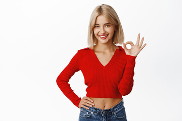 Okay, super good. Smiling fashionable blond girl showing ok, no problem, zero gesture and looking satisfied, standing over white background