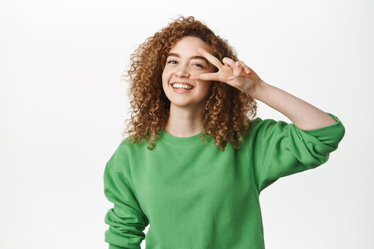 Happy smiling curly woman laughing, showing peace v-sign near eyes, cheerful upbeat emotions, posing against white background