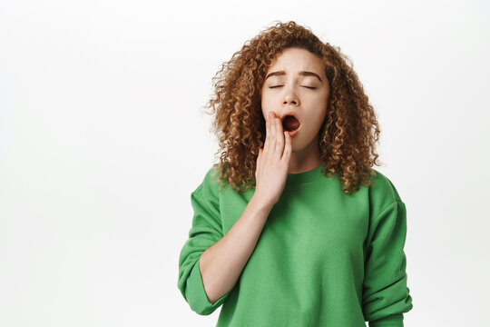 Portrait of tired curly girl, female student yawning with fatigue, feeling sleepy or bored, standing over white background, cover mouth with hand