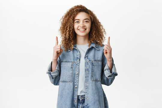 Portrait of smiling modern girl, millennial woman pointing fingers up on top, showing advertisement banner, standing against white background