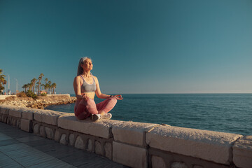 Fototapeta na wymiar Strong lady in sports clothes sitting by the beautiful seashore in lotus position and taining on sunny day