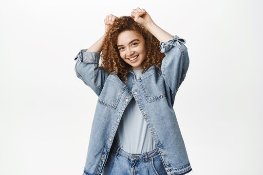 Image of cute stylish girl with curly hair, shows paws cuteness gesture and smiles, stands in casual clothes over white background