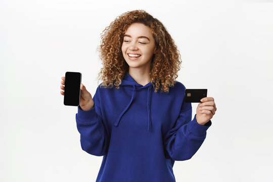 Mobile banking concept. Smiling young woman showing smartphone screen app, credit card, looking happy, recommending shopping application, white background