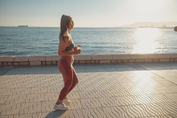 Fototapeta na wymiar Healthy sporty woman in sports top and leggings running by the seashore and looking at the sun