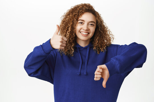 Choices. Portrait of young curly woman showing thumbs up and thumbs down, weighing variants, being indecisive, like or dislike, standing over white background