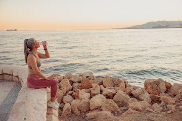 Asian lady in sportswear sitting on the edge of the coastal fence, drinking water, on background with blue sea and sunset
