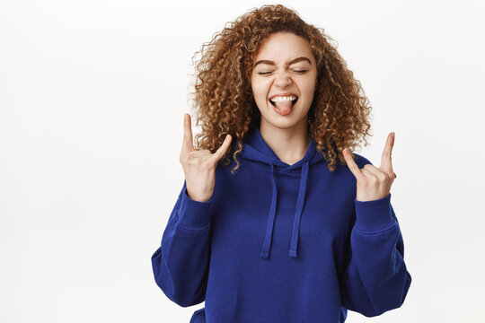 Portrait of sassy curly girl, showing rock on, heavy metal gesture and tongue, having fun, enjoying smth awesome, standing in blue hoodie over white background