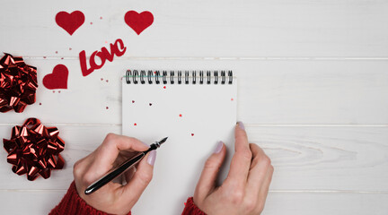 Woman's hands write a romantic congratulation to Valentine's Day in a notebook. Valentine's Day holiday concept. Top view. Copy space.