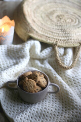 Bowl of chocolate chip cookies, soft knitted blanket, woven basket and lit candle on the table. Selective focus.