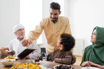 Happy muslim man pouring tea near interracial family and food at home.