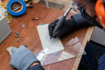 A craftsman wearing personal protective equipment does glass engraving with a rotary tool following a drawing on a paper