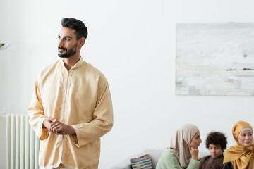 young muslim man looking away while standing near interracial family on blurred background.