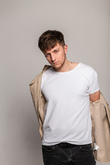 Cool stylish handsome young man with hairstyle in fashionable outerwear with white t-shirt puts on a beige jacket in the studio on a gray background