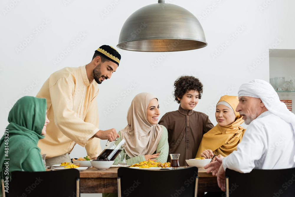 Wall mural young arabian man in skullcap pouring tea during dinner with interracial muslim family.