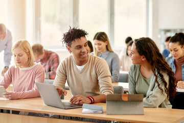 Happy African American student and his Caucasian classmate talk while learning on laptop at university classroom.