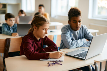 Caucasian schoolgirl takes notes while e-learning on computer with her African American classmate...