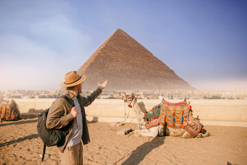 Tourist man in hat with camel background pyramid of Egyptian Giza, sunset Cairo, Egypt - obrazy, fototapety, plakaty