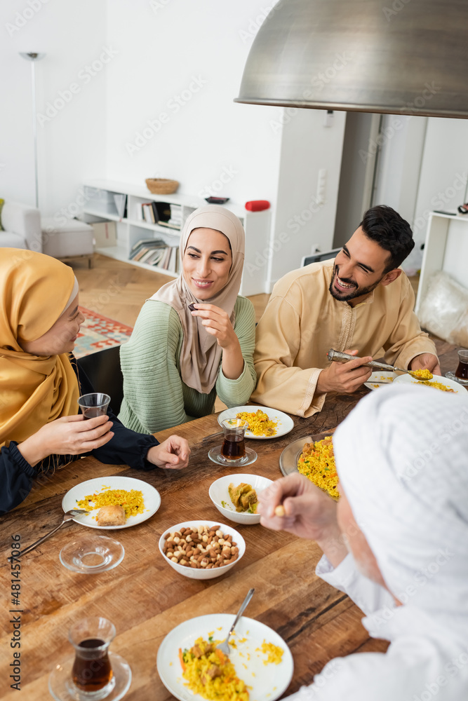 Wall mural mature asian woman holding glass of tea while talking to muslim family during dinner.