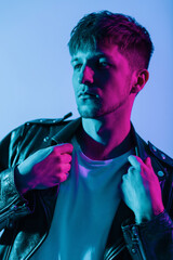 Colored fashion portrait of a handsome stylish guy with hairstyle in trendy outwear with vintage leather jacket on neon pink and purple light