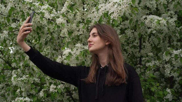 A smiling young girl takes pictures of herself in the park against the background of a blooming white apple tree. Teen takes selfie on smartphone during wind. High quality 4k footage