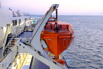 Red lifeboat hangs on ferry. White deck of large passenger ferry.  Lights on deck in morning. 