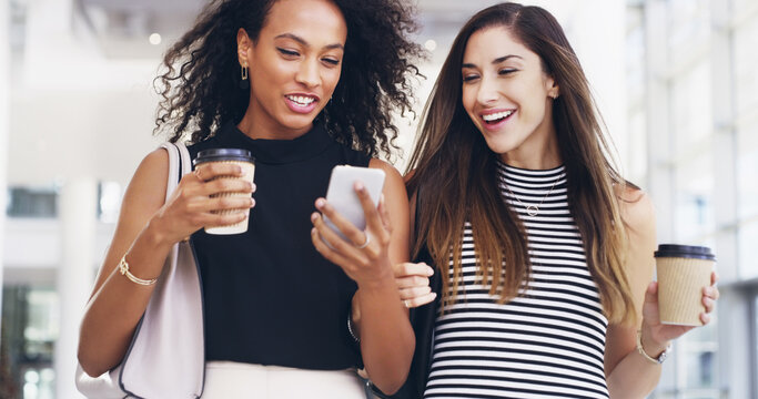 They share everything. Cropped shot of two young businesswomen chatting and using a smartphone while walking in an office on a coffee break.