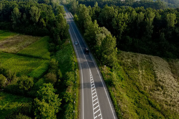 Aerial view of cars driving along the empty roadriding along trees and fields on sunny morning.Travelling, car rental, insurance concept