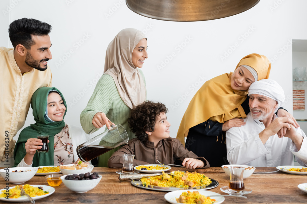 Wall mural cheerful arabian woman pouring tea during dinner with multiethnic muslim family.