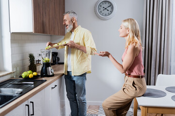 Side view of smiling woman sitting near husband preparing smoothie in kitchen.