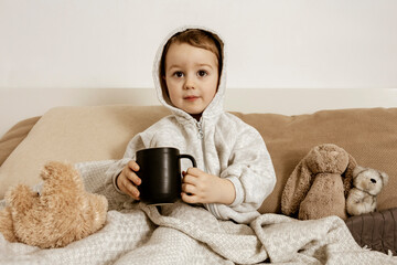 Sick little boy drinking hot tea on the bed at home. Unwell, illness child wrapped in a blanket,...