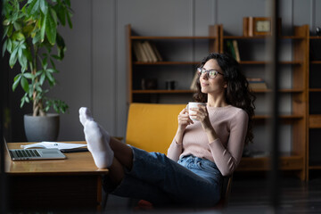 Wellbeing at work. Young relaxed female employee resting at workplace with cup of coffee and legs...