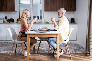 Side view of positive mature couple holding cups near pie in kitchen.