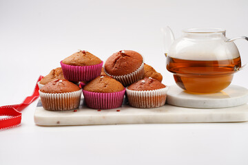 Several homemade white and pink muffins on marble board on white surface, red ribbon, heart-shaped red and white sugar decoration and tea-pot