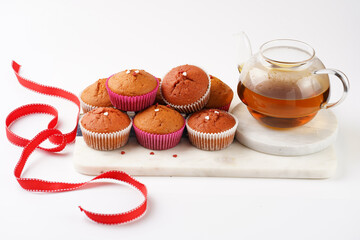 Several homemade white and pink muffins on marble board on white surface, red ribbon, heart-shaped red and white sugar decoration and tea-pot