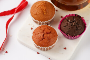 Several homemade white and pink muffins on marble board on white surface, red ribbon, heart-shaped red and white sugar decoration and tea-pot
