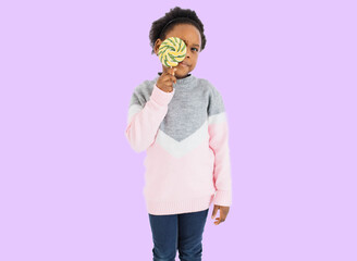 African little child using sweet lollipop for hiding one eye in studio. Cute female kid taking fun with delicious candy in hands over lilac background.
