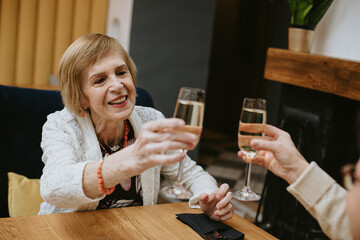 Smiling elderly fair-haired woman with makeup and orange bracelet dressed in white jacket clinking glasses of wine or champagne in cafe celebrating her meeting with friends