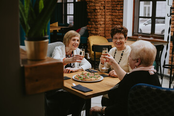 Smiling elderly dark-haired and fair-haired women with makeup dressed in white jackets taking glasses of wine or champagne in cafe celebrating their meeting with friends